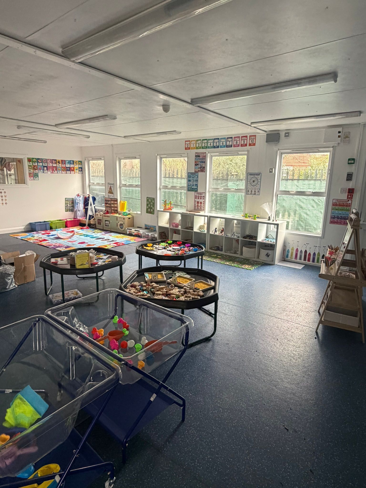 A well-lit, empty preschool classroom with tables, cubbies, play materials, and large windows along the back wall.