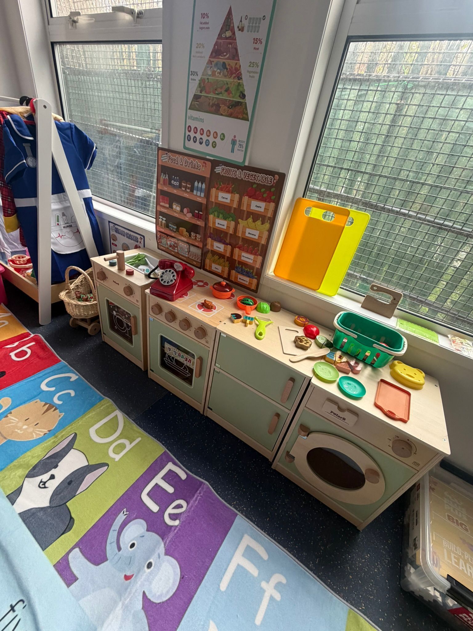 A play kitchen and alphabet rug in a classroom, featuring wooden cabinets, a sink, and educational wall posters.
