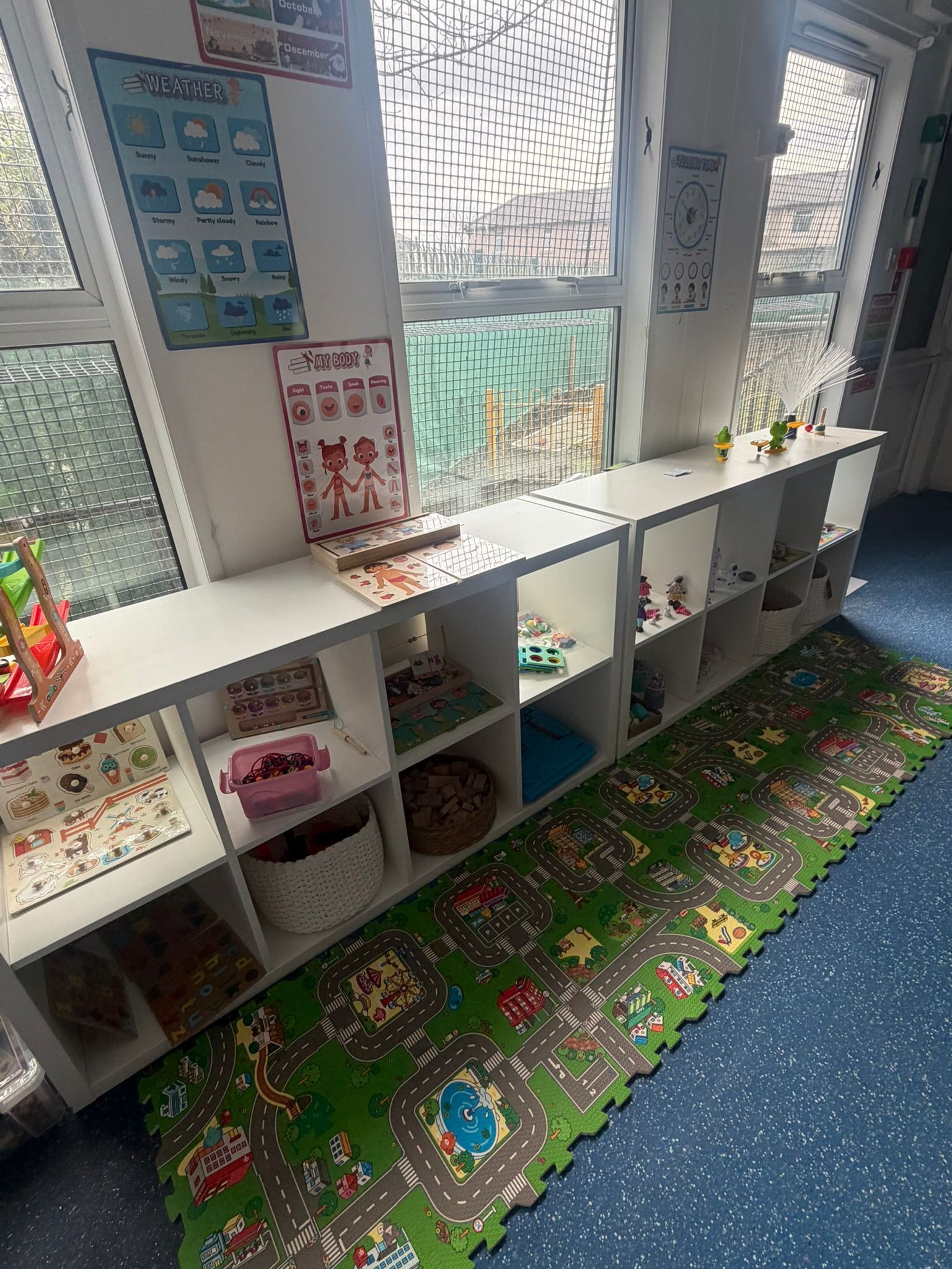 A low, white grid shelving unit in a classroom, filled with toys and games, sits on a colorful play mat featuring roads.