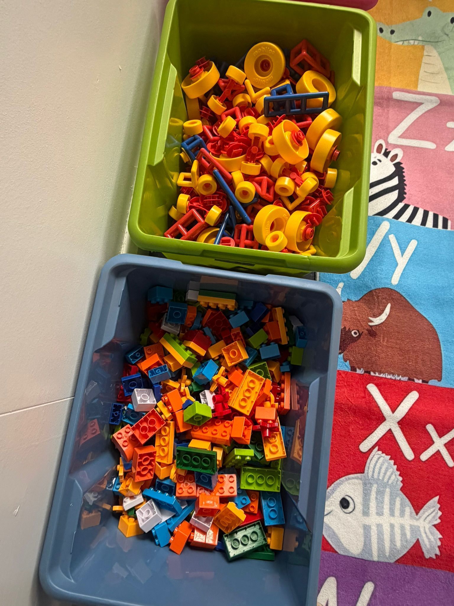 Two plastic storage bins filled with colorful building blocks sit on a children’s alphabet play mat.