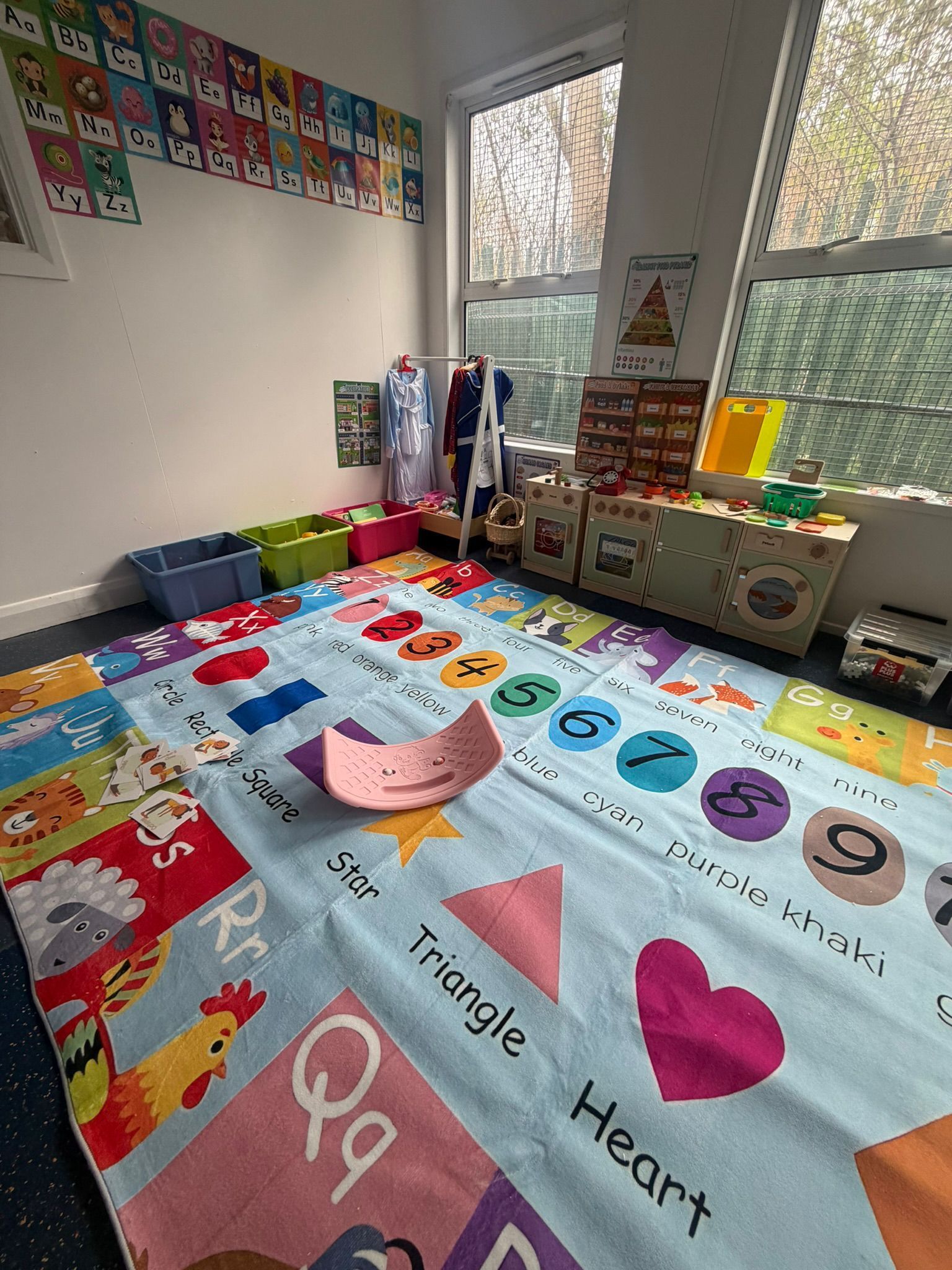A colorful playroom rug featuring letters, shapes, and numbers, with a wooden curved balance board sitting on top.