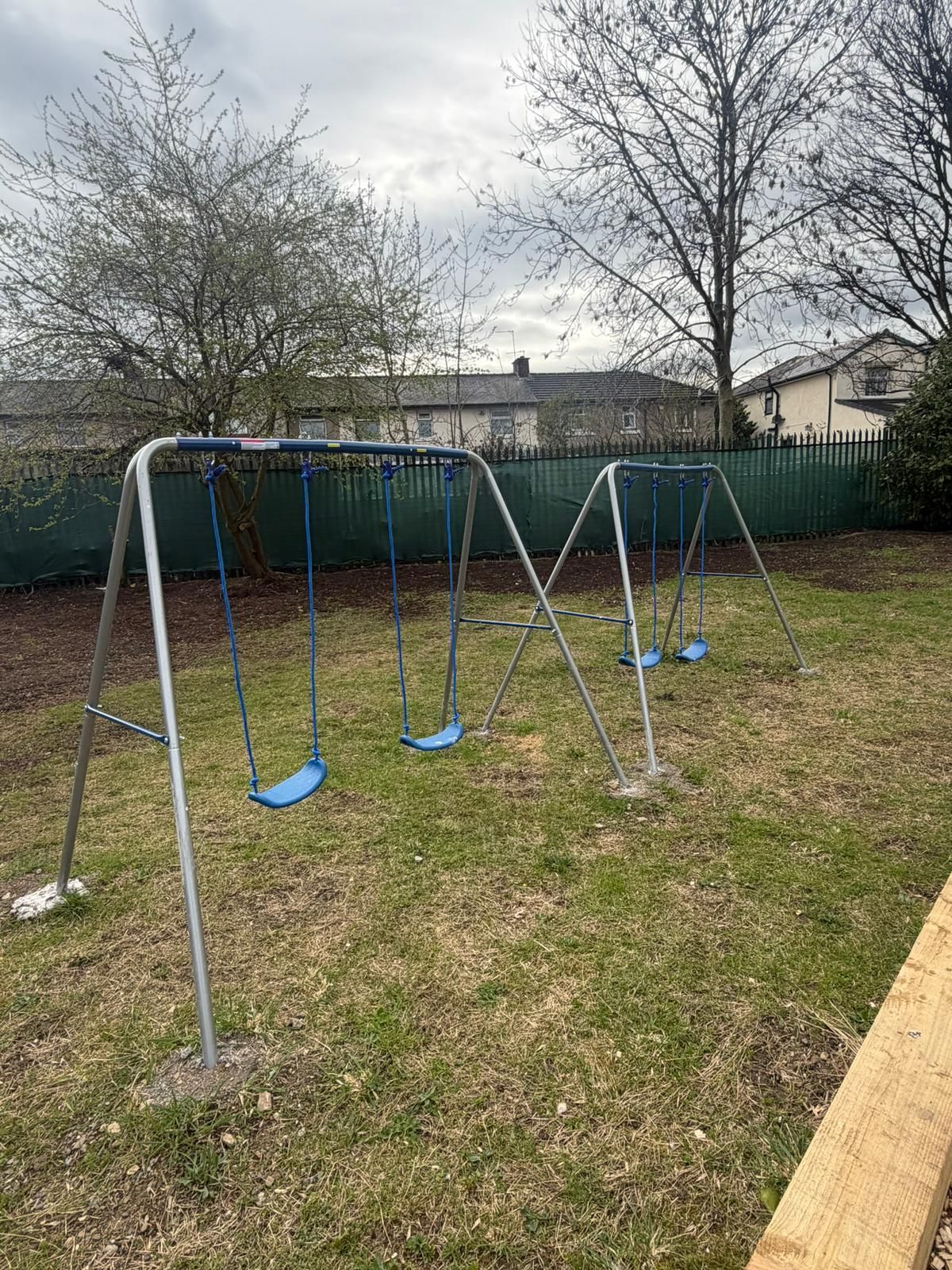 A metal swing set with four blue swings stands on a grassy backyard lawn in front of a green privacy fence and trees.
