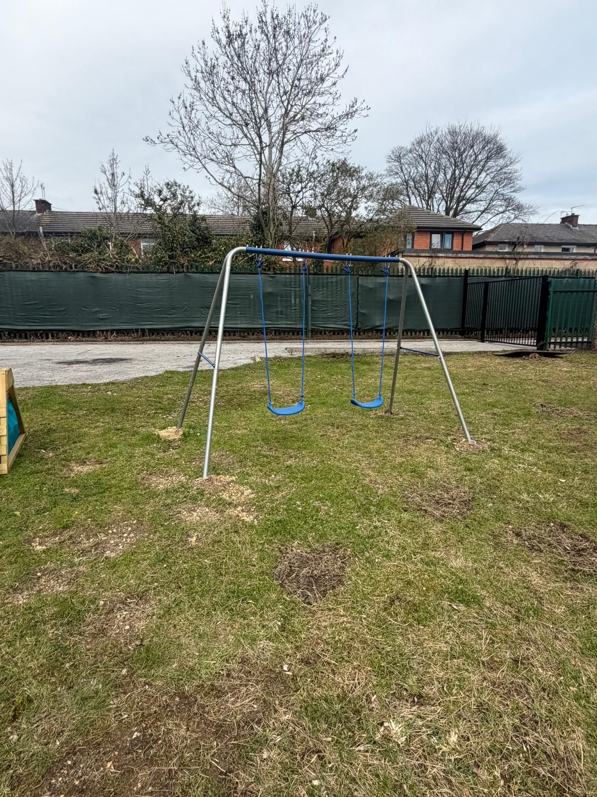 A metal swing set with two blue seats stands in a grassy yard, backed by a tall green fence and trees.
