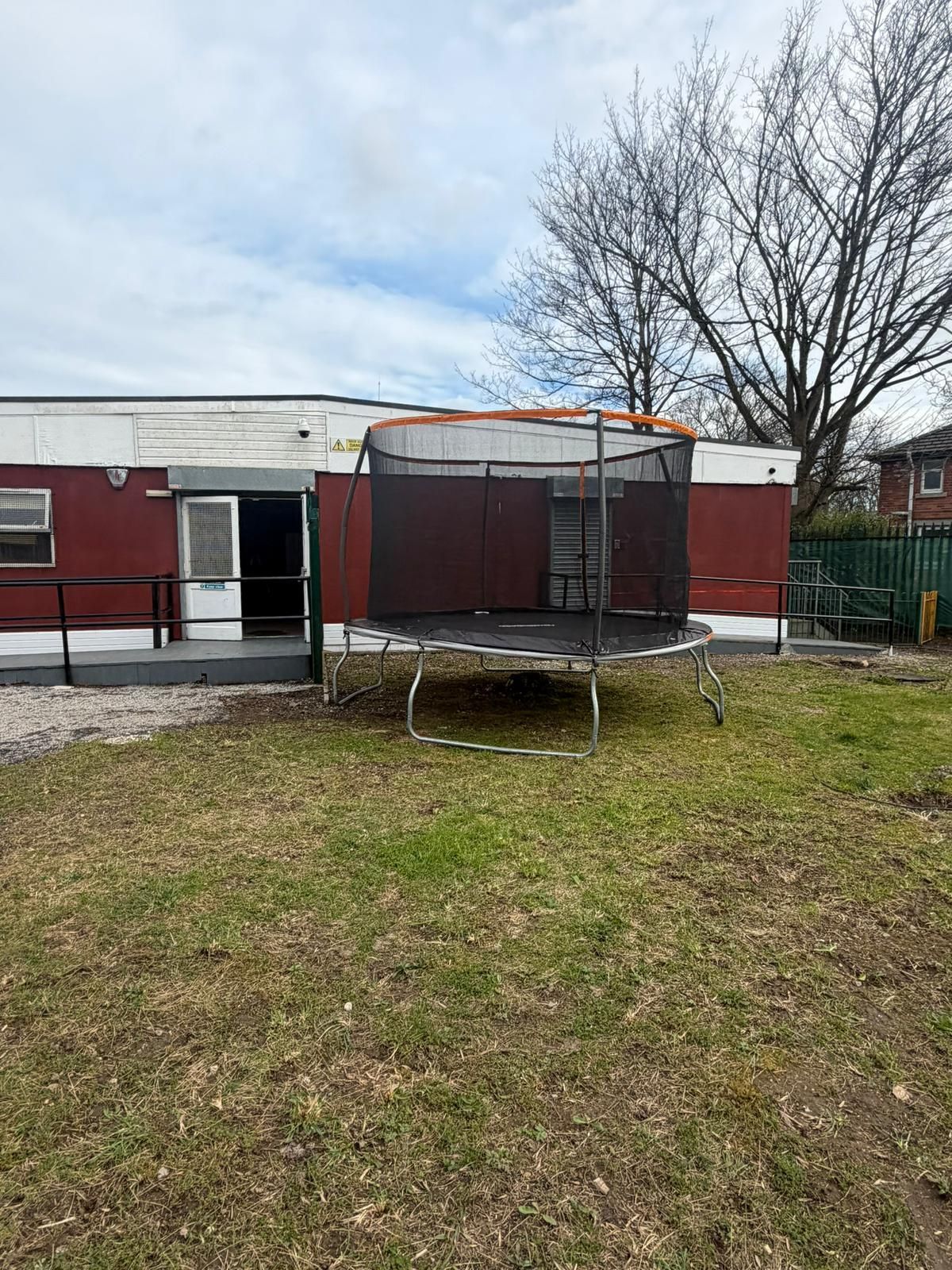 A large circular trampoline sits in a grassy yard in front of a single-story maroon building with a white upper section.