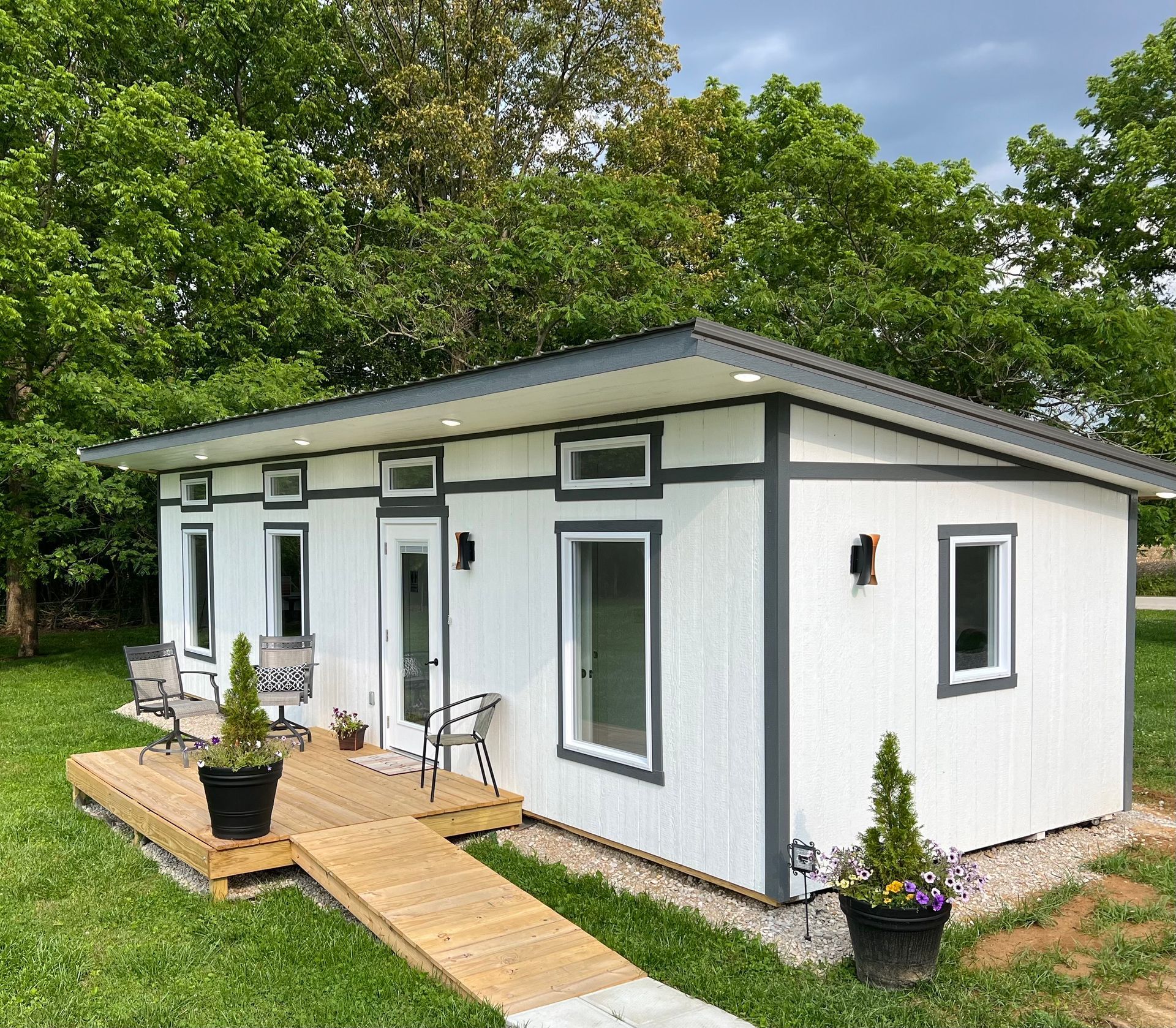 White tiny house with a gray roof and trim, a ramp, and a small deck, set in a grassy area.
