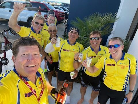 Group of cyclists in yellow jerseys smile, raising drinks and posing outdoors near vehicles.