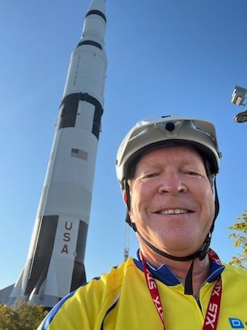 Man in cycling gear smiles in front of a large Saturn V rocket at a museum; blue sky background.