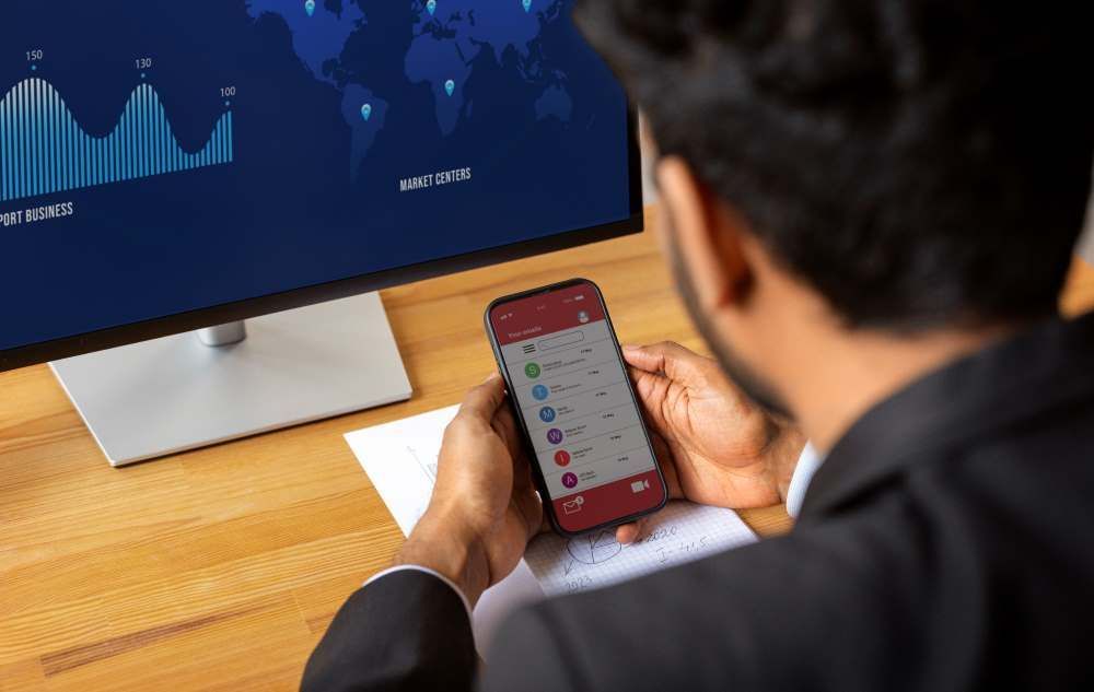 Um homem está sentado em uma mesa segurando um celular em frente a um monitor de computador.