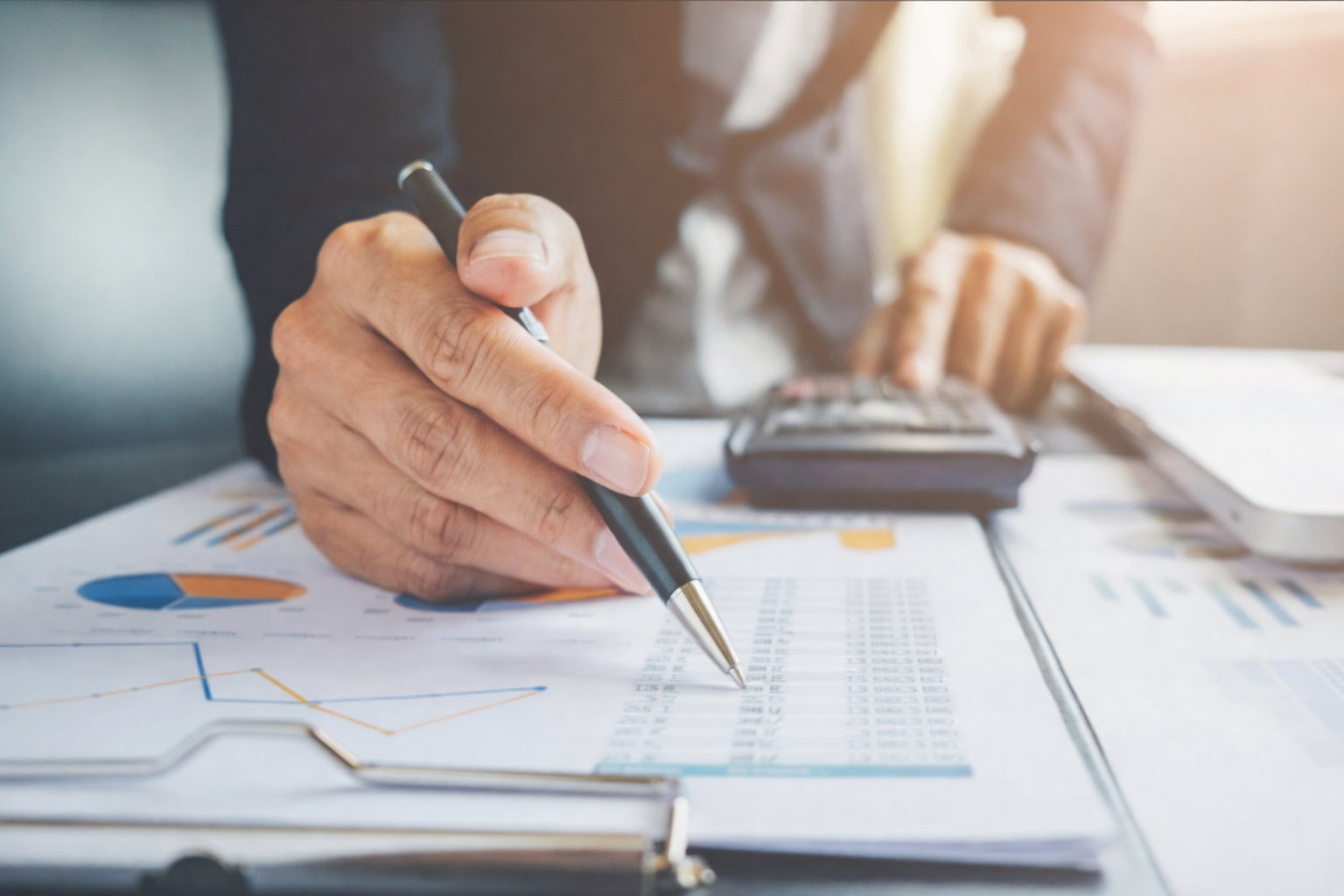 Pessoa de terno usando caneta, calculadora e relatórios financeiros.