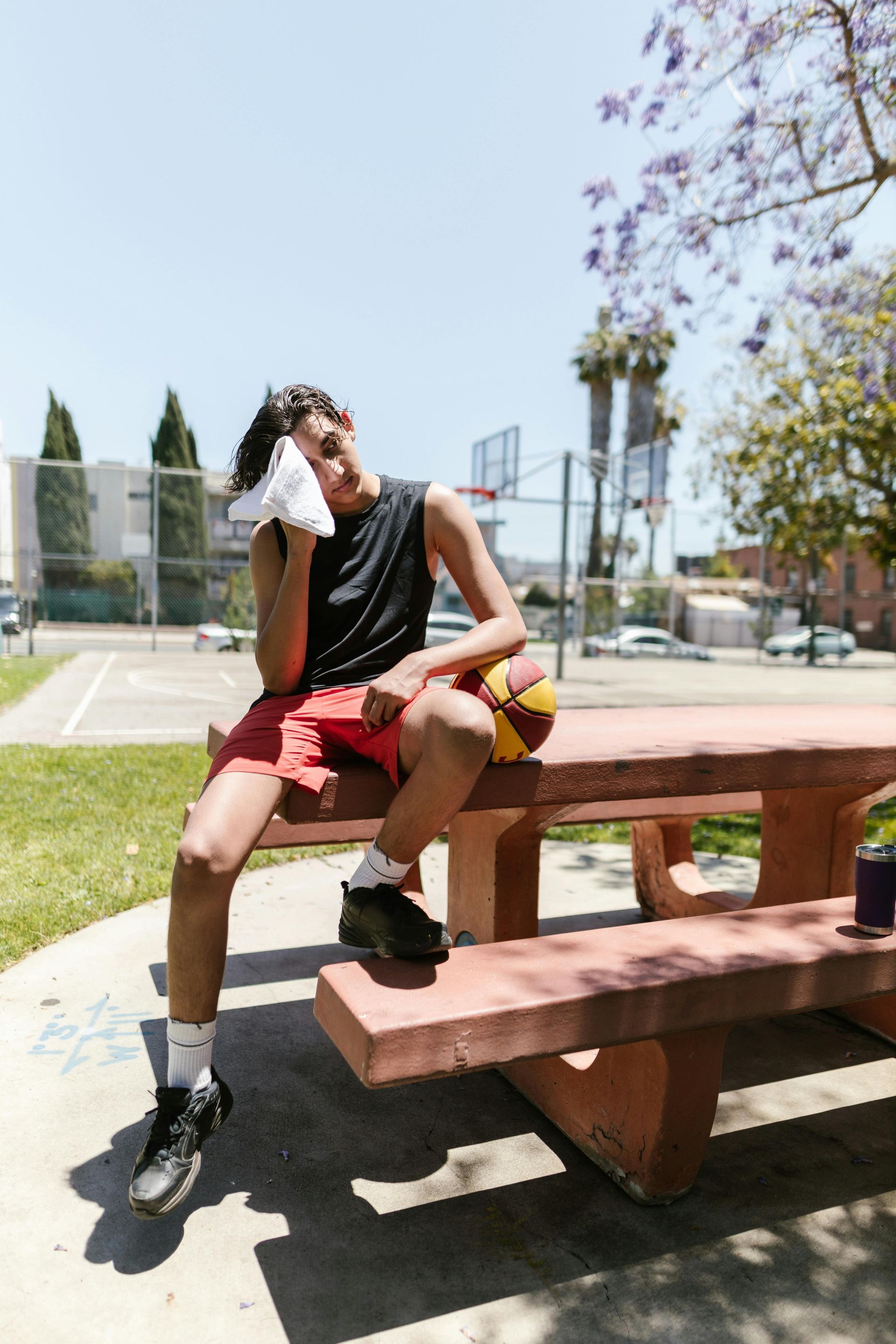 A young person wipes sweat with a towel, resting on a picnic table with a basketball in a sunny park.