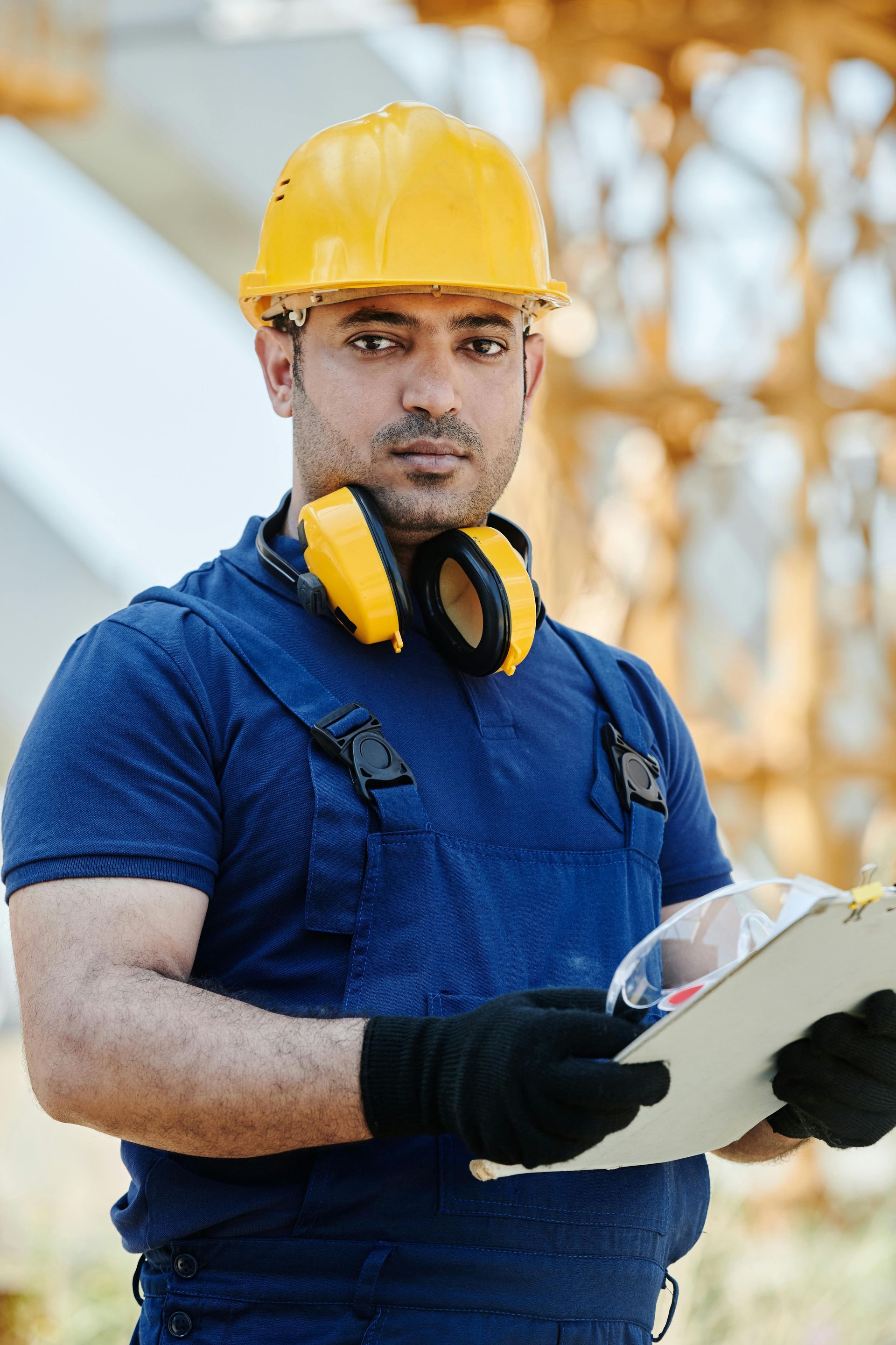 Construction worker in yellow hard hat and blue uniform holding a clipboard, with ear protection around his neck.