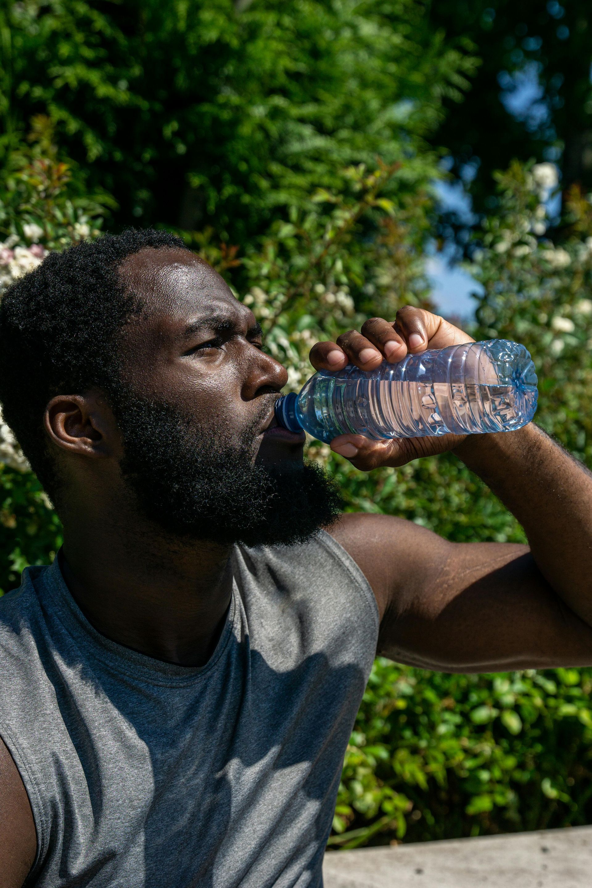 Sweaty Black man drinking water from a clear plastic bottle outdoors, with green foliage in the background.