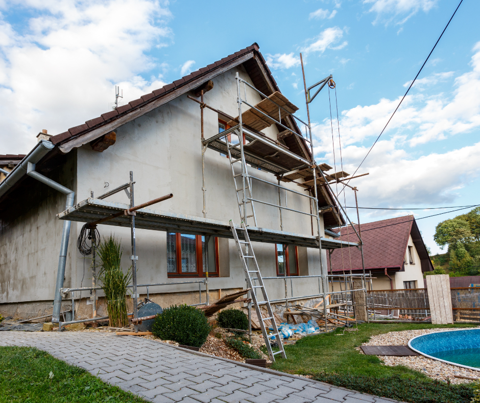 House exterior undergoing renovations, with scaffolding. Brown roof, white siding, blue sky, and a pool.