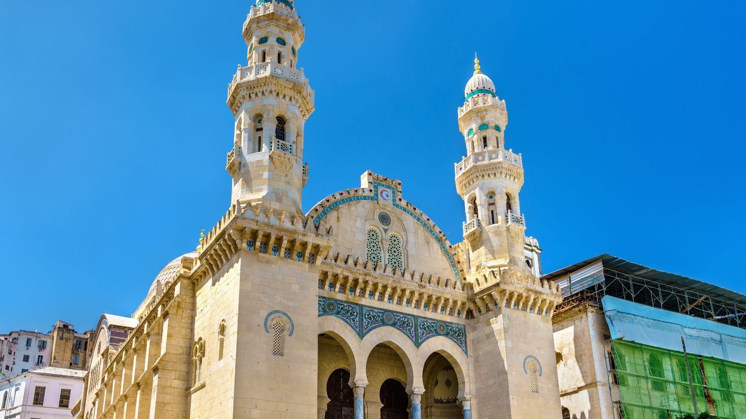 A historic mosque with two ornate minarets and intricate stone arches stands under a vibrant blue sky in Algiers.