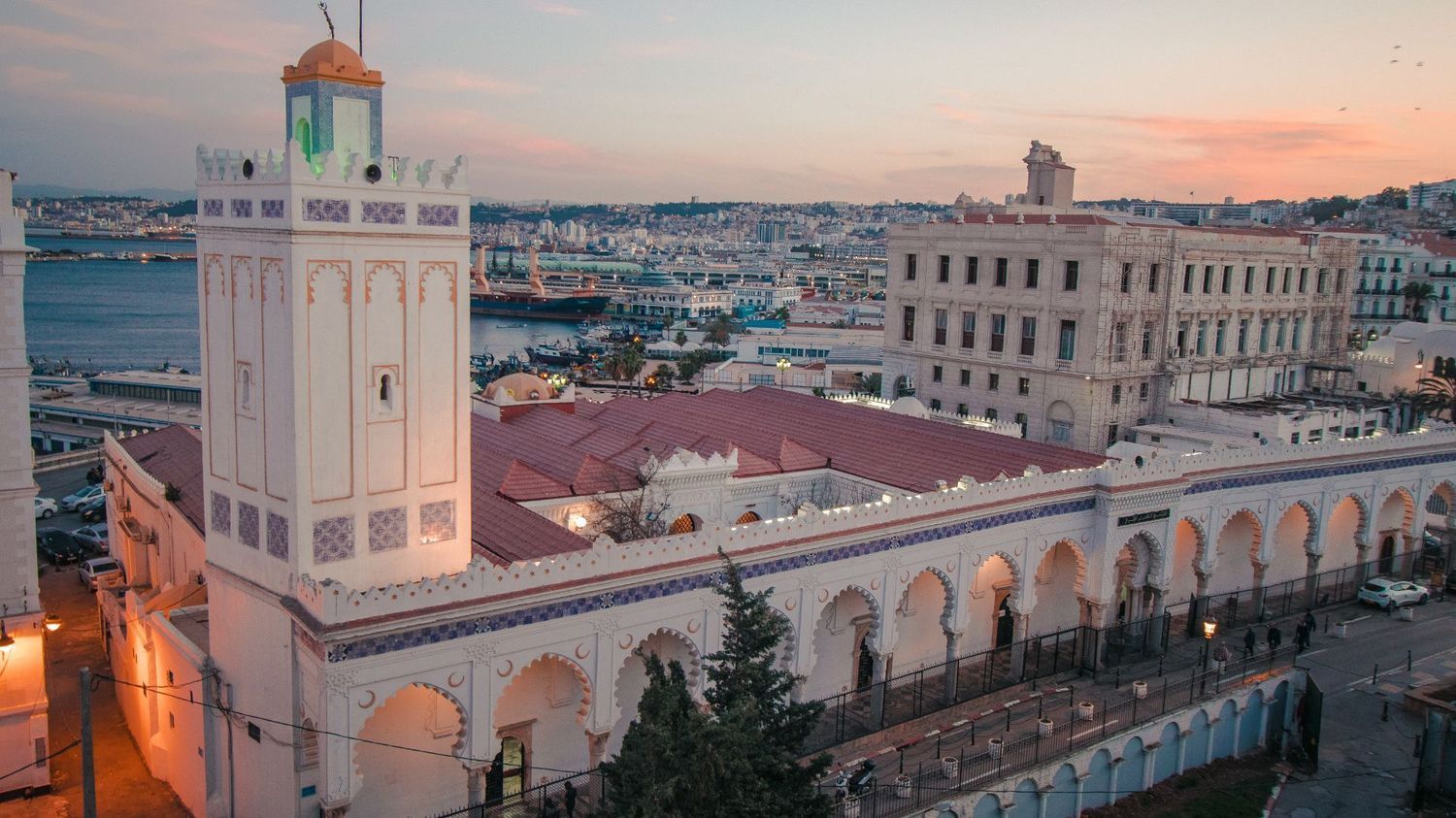 Aerial view of the Ketchaoua Mosque in Algiers, featuring a white minaret, red roof, and arched portico at sunset.