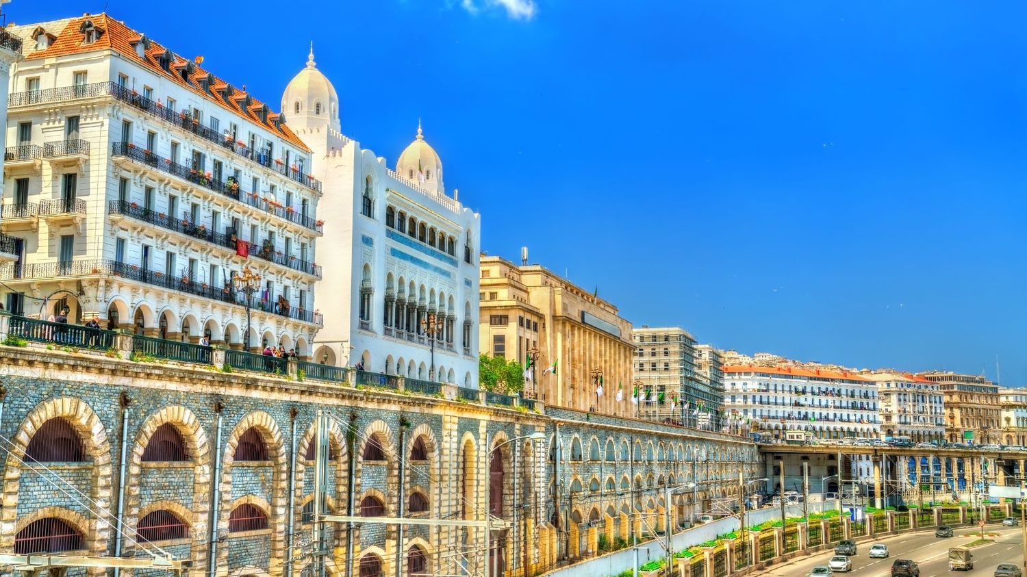 Historic white buildings with domed roofs stand above an arched stone retaining wall in Algiers under a clear blue sky.