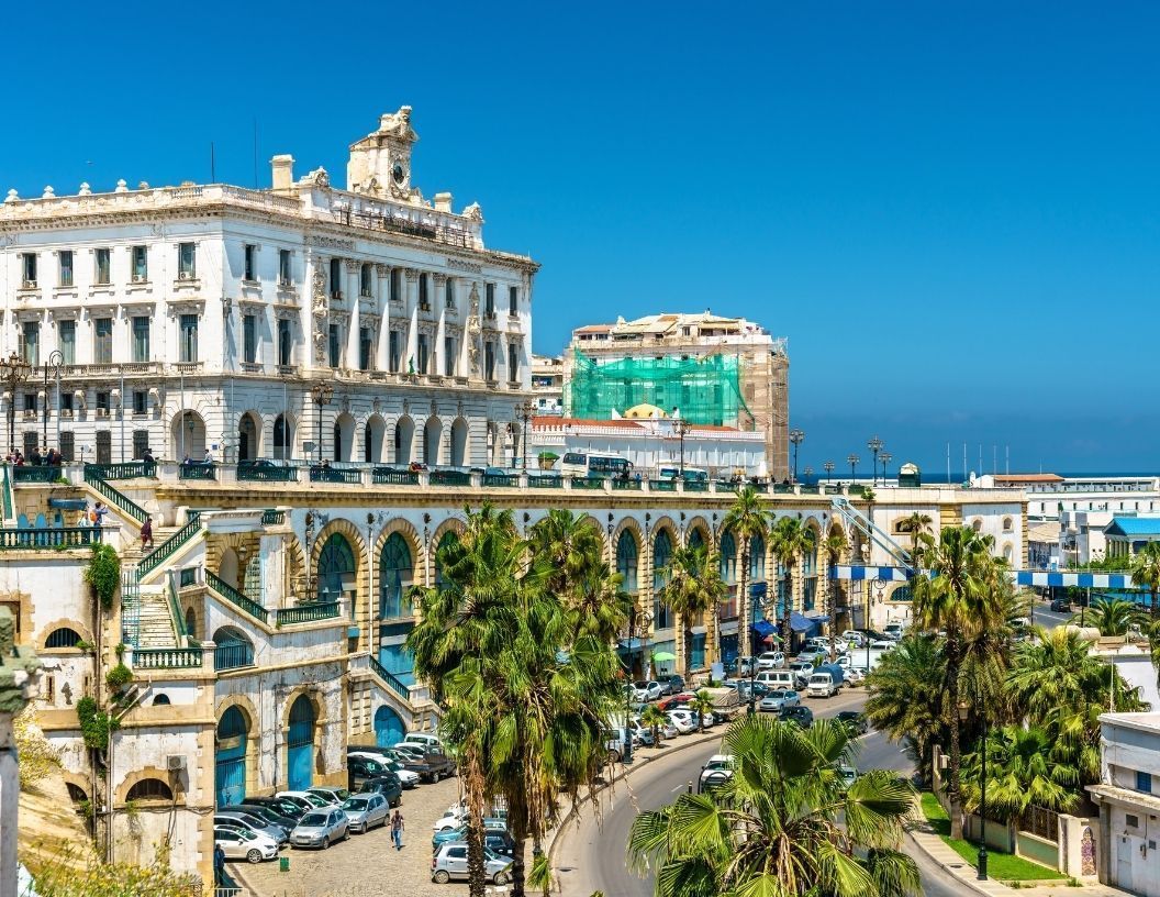 The white, neoclassical facade of the Palais du Gouvernement with arched walkways above a palm-lined street in Algiers.