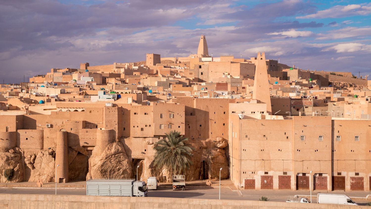 An ancient, earth-toned desert city built on a rocky hill with a prominent minaret under a cloudy sky.