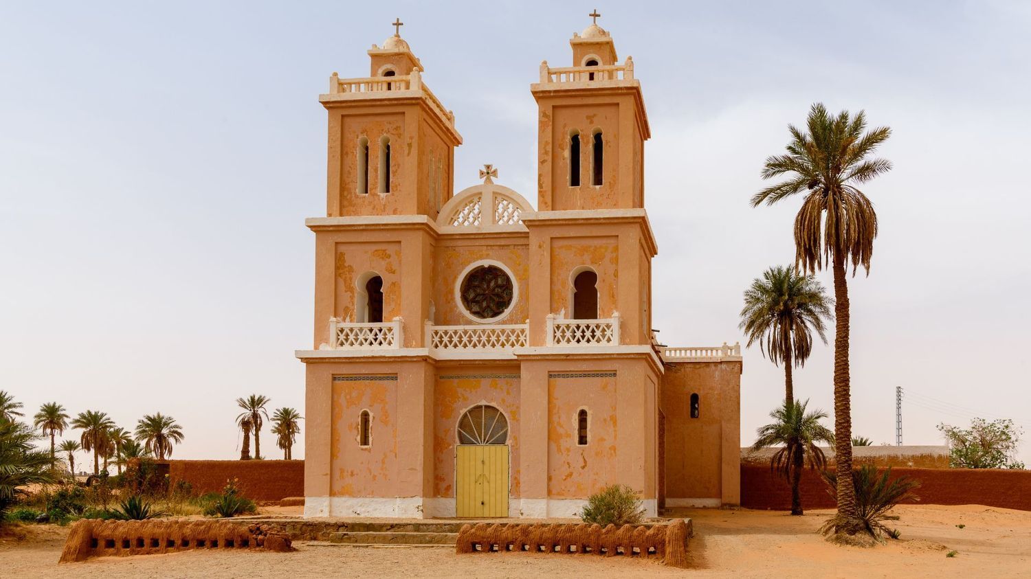 A tan, two-towered church with arched windows and a yellow door stands in a desert landscape with palm trees.