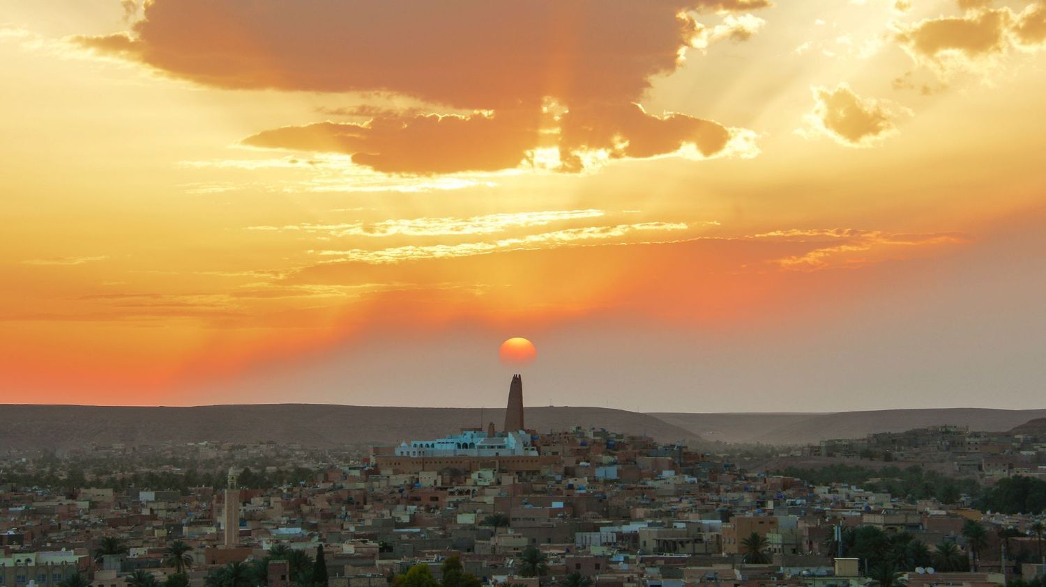 A sunset casts a golden glow over a dense hillside town with a tall, central tower silhouetted against the bright sun.