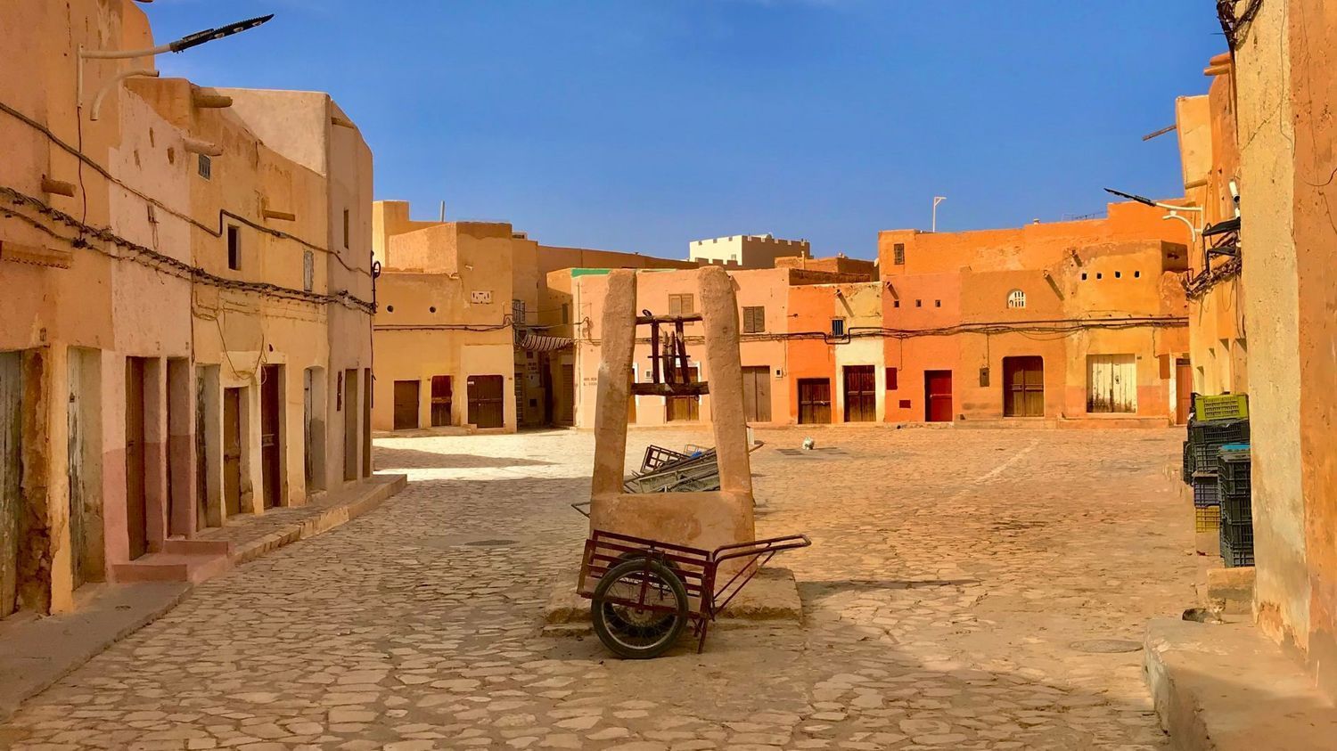 A sunny, orange-hued stone square in an old town, featuring a central stone well and a wooden cart on cobblestones.