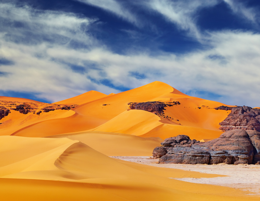 Golden sand dunes under a vibrant blue sky with wispy clouds, featuring rocky outcrops in the foreground.