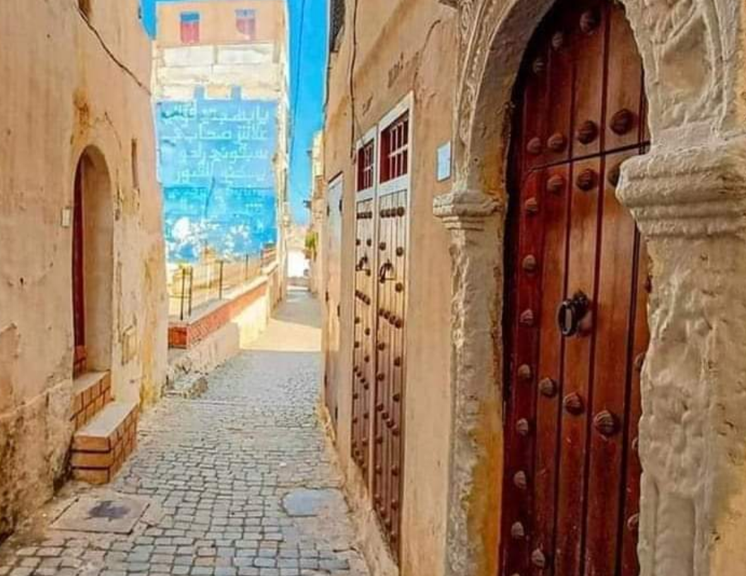 A cobblestone alleyway in a historic town featuring rustic tan buildings, carved wooden doors, and a bright blue wall.
