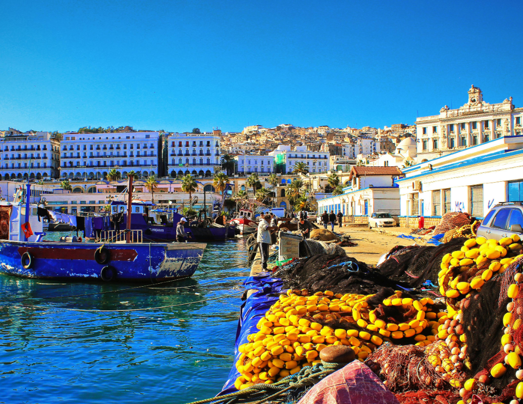 A blue fishing boat at a harbor quay with piles of yellow fishing nets in the foreground and city buildings behind.