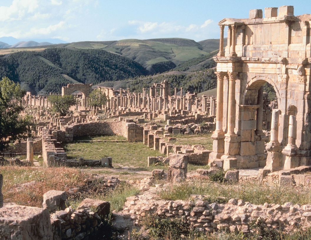 Ancient Roman ruins in Timgad, Algeria, featuring a stone triumphal arch in the foreground and columns against hills.