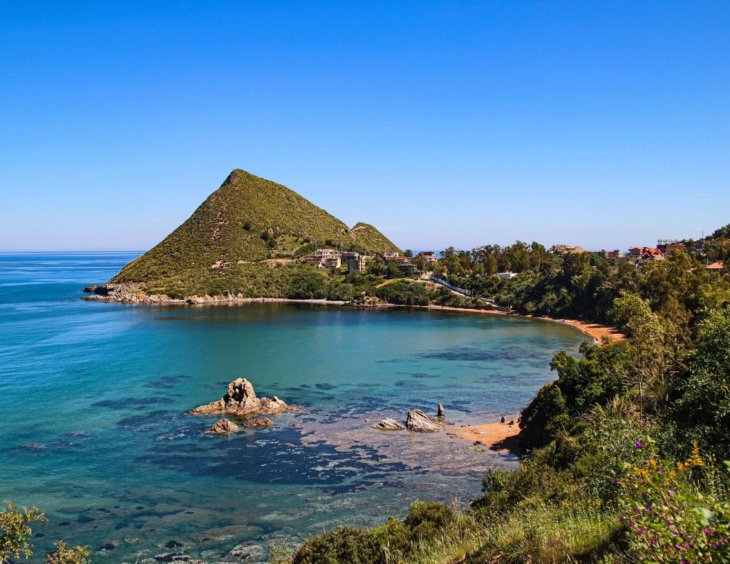 A scenic view of a triangular green hill overlooking a turquoise bay with rocky formations and a small, sandy beach.