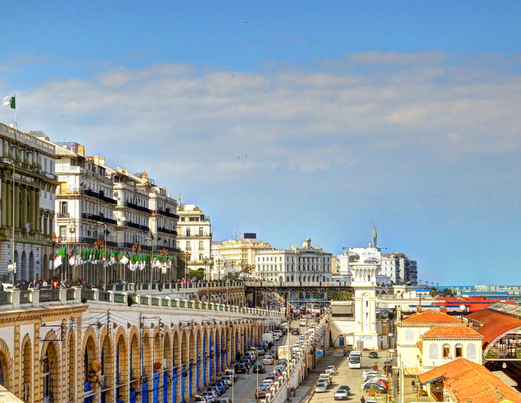 A scenic view of the Algiers waterfront, featuring ornate white arched buildings along a road near the Mediterranean Sea.