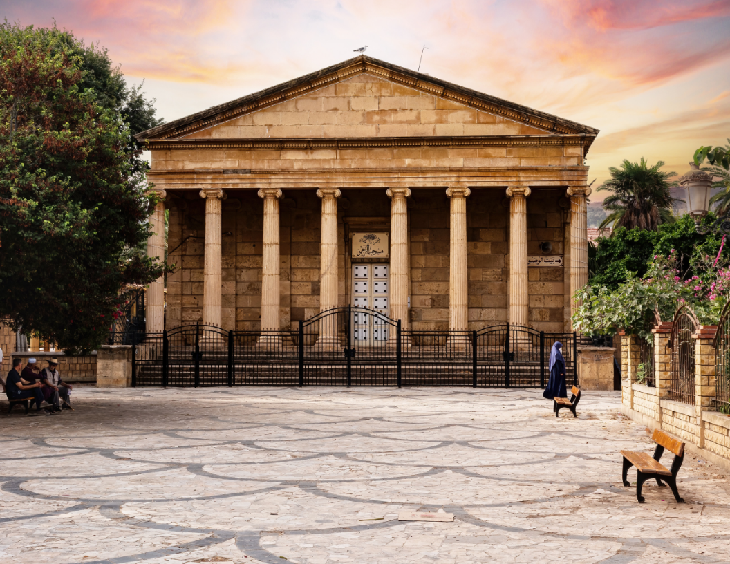 A classical-style stone building with a columned porch, set behind an iron fence in a cobblestone courtyard at sunset.