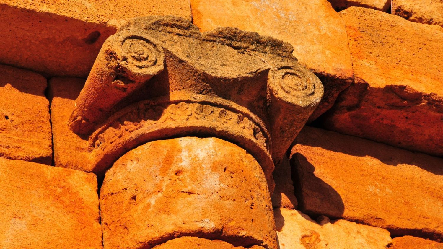A weathered, orange sandstone Ionic capital sits atop a column against a background of stacked, matching brickwork.
