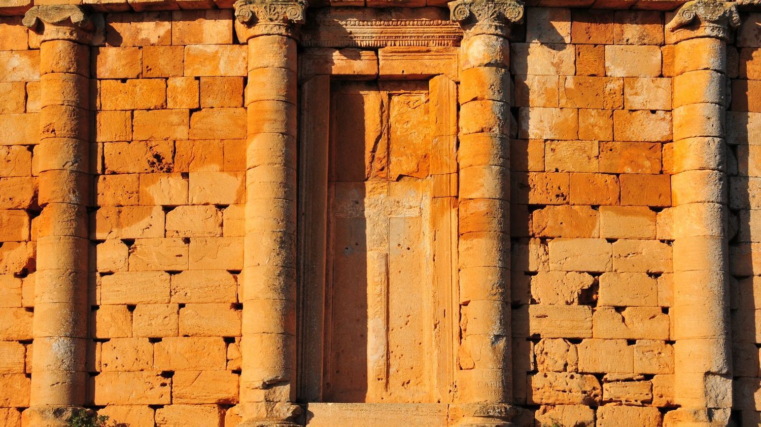 A stone wall featuring three vertical columns flanking a tall, narrow, recessed rectangular doorway in golden sunlight.