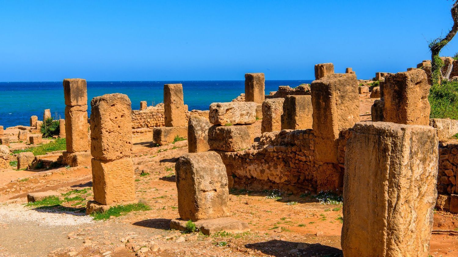 Ancient stone ruins with rectangular columns overlooking a bright blue sea under a clear sky.