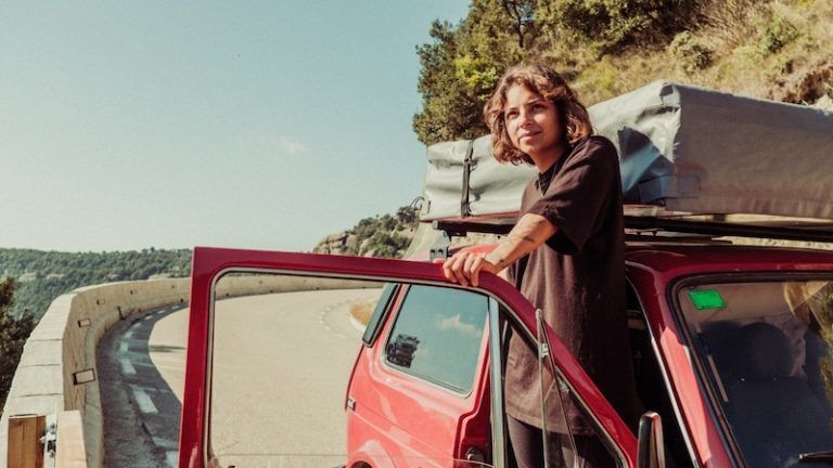 A person in a black shirt stands by the open door of a red car, parked on a winding mountain road with gear on the roof.
