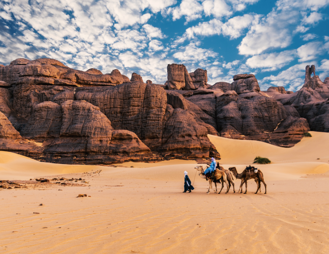 A person walks beside two camels near tall, dark sandstone rock formations in a vast, sunny desert under a cloudy sky.