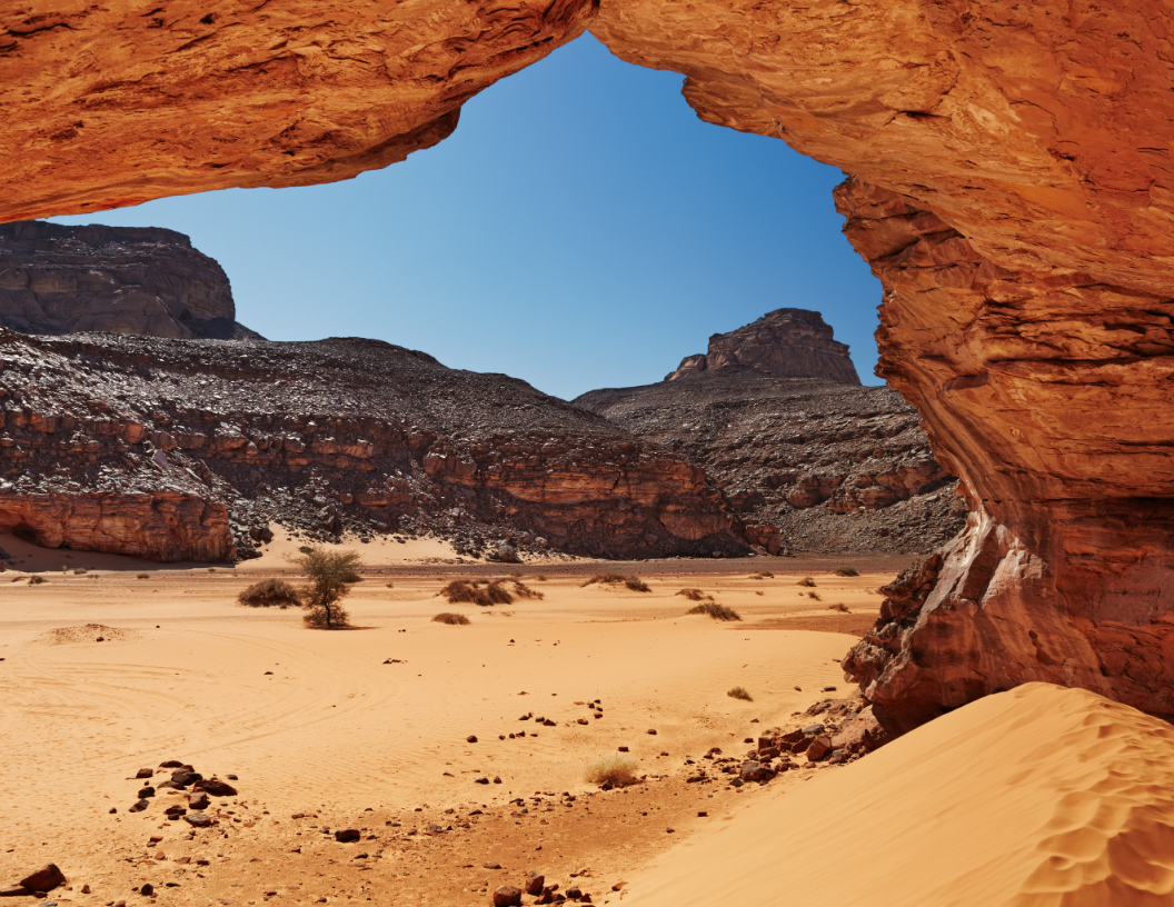 A natural sandstone arch frames a view of a vast, arid desert landscape with rugged mountains under a clear blue sky.