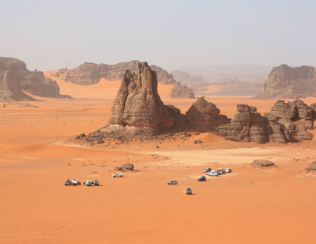 Off-road vehicles parked on orange sand dunes near towering, dark rock formations in a desert landscape.