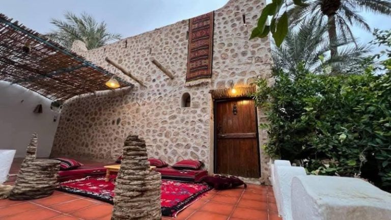 A patio with a stone wall, red carpet, floor cushions, wooden door, and palm trees in a rustic desert-style setting.