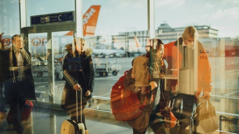 People with luggage walk past a window in an airport terminal, with an orange EasyJet plane visible on the tarmac.