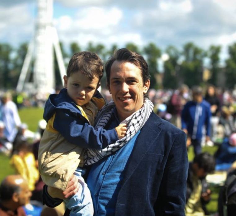 A person holding a young child in their arms, both smiling outdoors with a tall white structure in the blurred background.