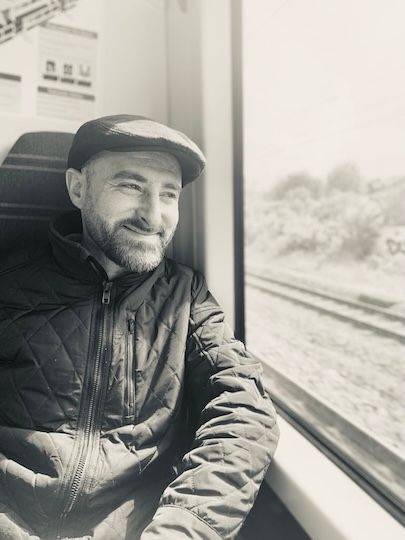 A person wearing a flat cap and a quilted jacket looks out a train window while smiling slightly.