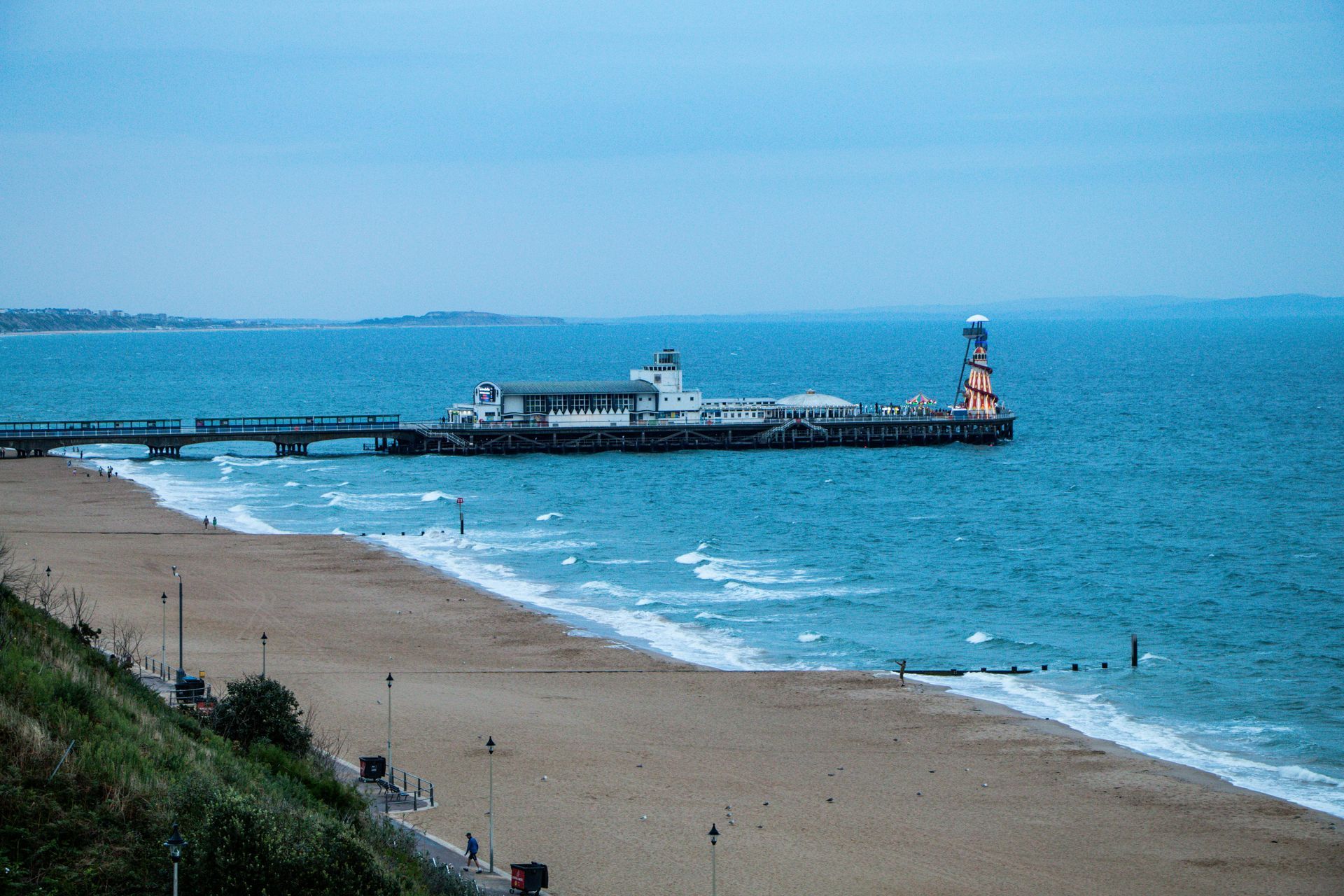 Boscombe Pier stretching into a blue sea, with a beach and sand cliffs in the foreground under a clear sky.