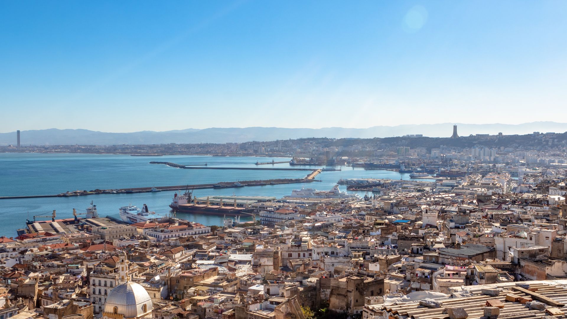 A high-angle view of Algiers, showing the dense white buildings of the Casbah, the port, and the Mediterranean Sea.