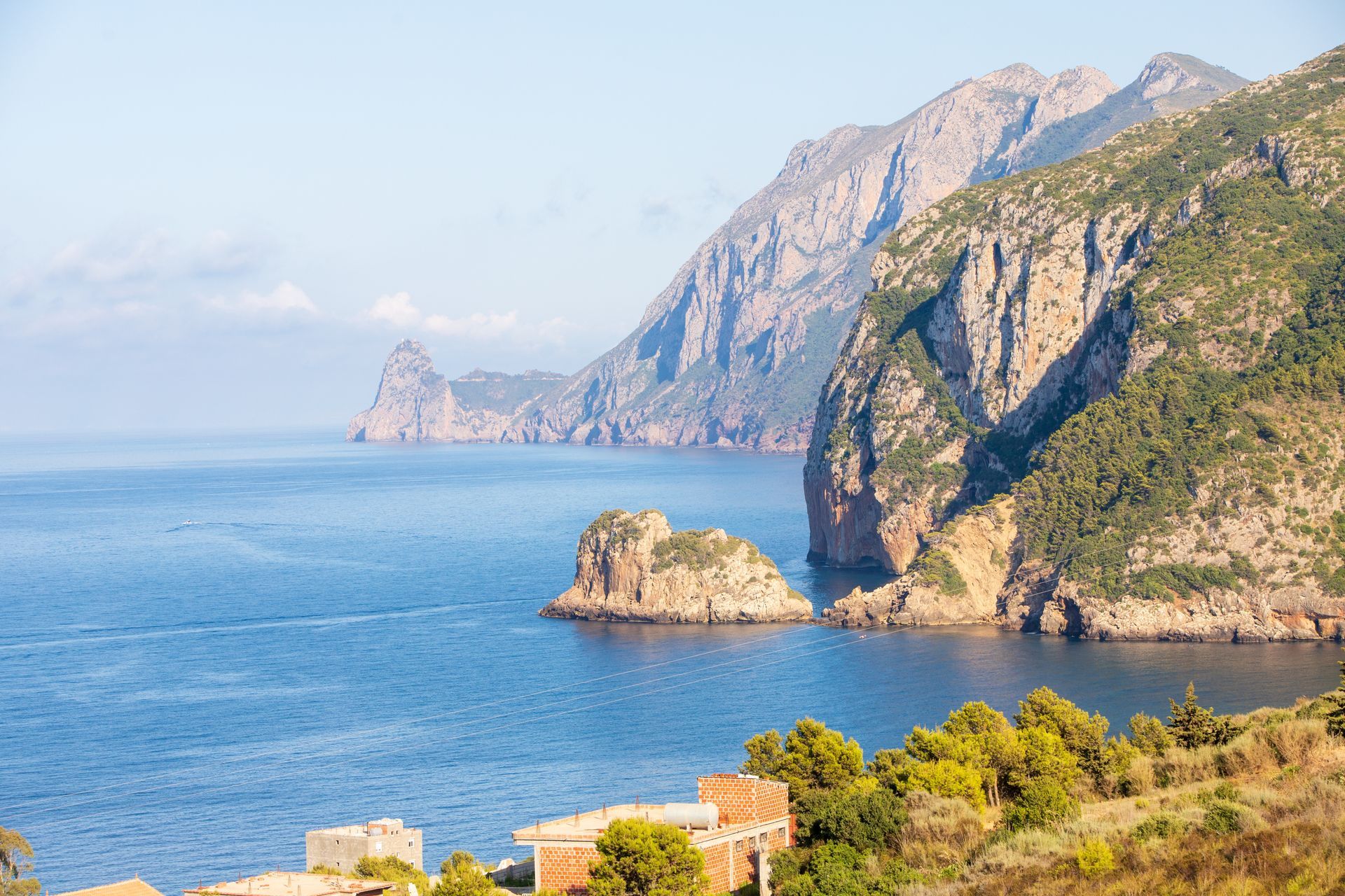 Rugged cliffs and a small rocky island jut into the deep blue Mediterranean sea under a clear, bright sky.