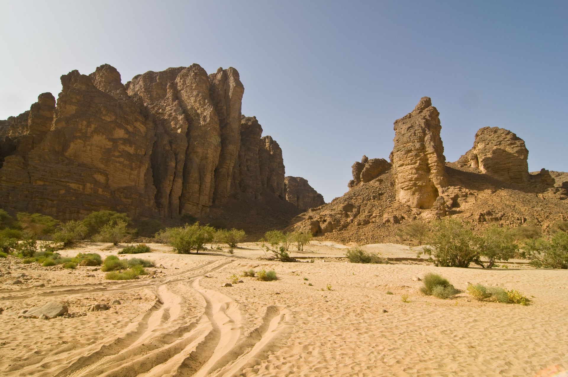 Sandy desert tracks lead toward towering, weathered rock formations under a clear blue sky.