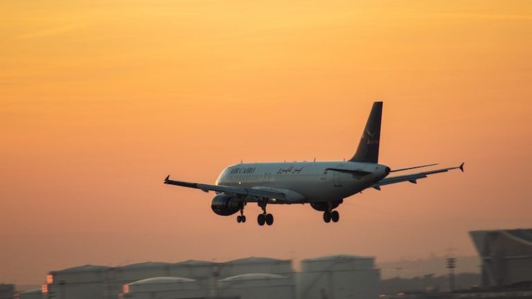 A commercial airplane in landing configuration flies low against a warm, orange sunset sky above industrial structures.