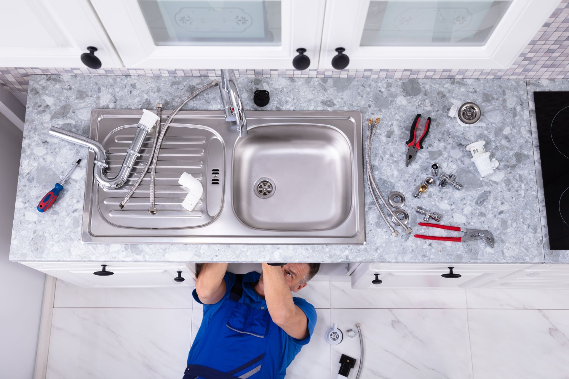 Overhead view of a plumber working under a kitchen sink with tools spread on the counter.