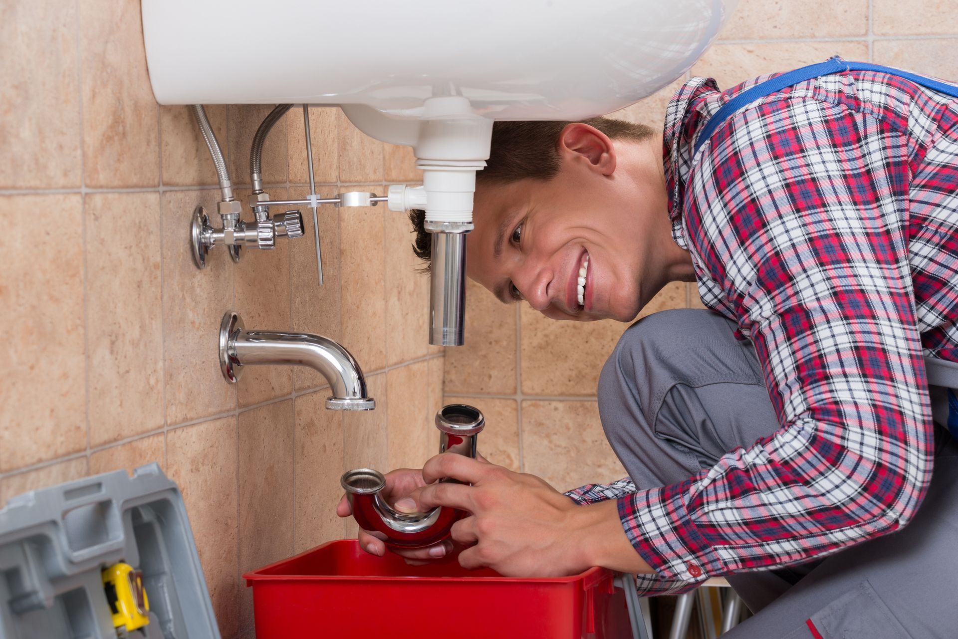 A smiling male plumber installs a silver sink pipe inside a bathroom, crouching in front of it.